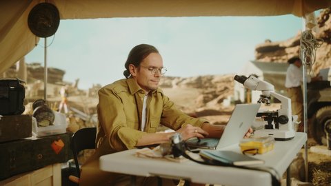 Archaeological Digging Site: Smart Male Archaeologist Doing Research, Using Laptop, Analysing Fossil Remains, Ancient Civilization Culture Artifacts. Team of Historians work on Excavation Site