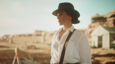 Portrait of Beautiful Female Adventurer Wearing a Hat Posing and Looking at Camera. Stylish Great Archaeologist Standing with Ancient Civilization, Fossil Remains Archeological Site in Background