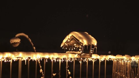 Night view of birdhouse and feeder covered by snow with glowing christmas light. Snowflakes are falling from top. Fairytale scene with bird wooden house, snow and ice in winter.