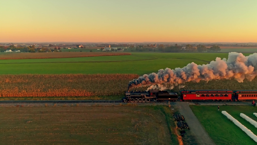 Aerial View of a Steam Engine with Passenger Cars at the Golden Sunrise Approaching with a Full Head of Steam and Smoke Traveling Thru the Farmlands
 Royalty-Free Stock Footage #1067288167