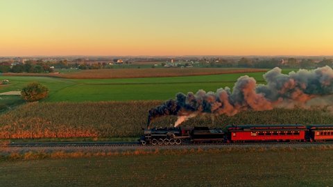 Aerial View of a Steam Engine with Passenger Cars at the Golden Sunrise Approaching with a Full Head of Steam and Smoke Traveling Thru the Farmlands
