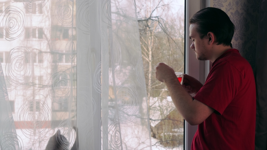 A young guy stands by the window with a cup of black tea in his hands and looks at the snowy street, and a small gray kitten sits on the windowsill
