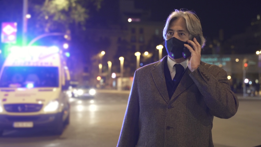 Man with face mask on phone call in front of Ambulance flashing lights in the city at night. Businessman Calling for help. Accident Urgency during Coronavirus Pandemic