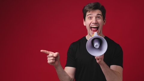 Cheerful shocked young man 20s years old in casual black t-shirt posing isolated on red background studio. People sincere emotions lifestyle concept. Pointing index finger aside screaming in megaphone