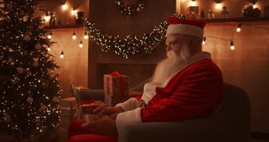 Santa Claus sits next to a Christmas tree on a comfortable soft sofa, holds a laptop in his lap and answers emails from young children.