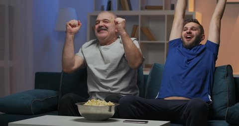 Father and son are watching a sports match on TV while sitting on the couch at home. Root for favorite team. They raise their hands enthusiastically and shout after a goal has been scored.
