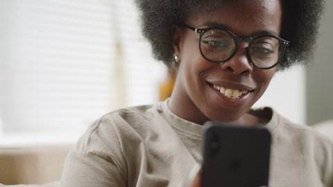 Close up shot of young positive Afro-American woman smiling and chatting via video call on smartphone while sitting on sofa at home