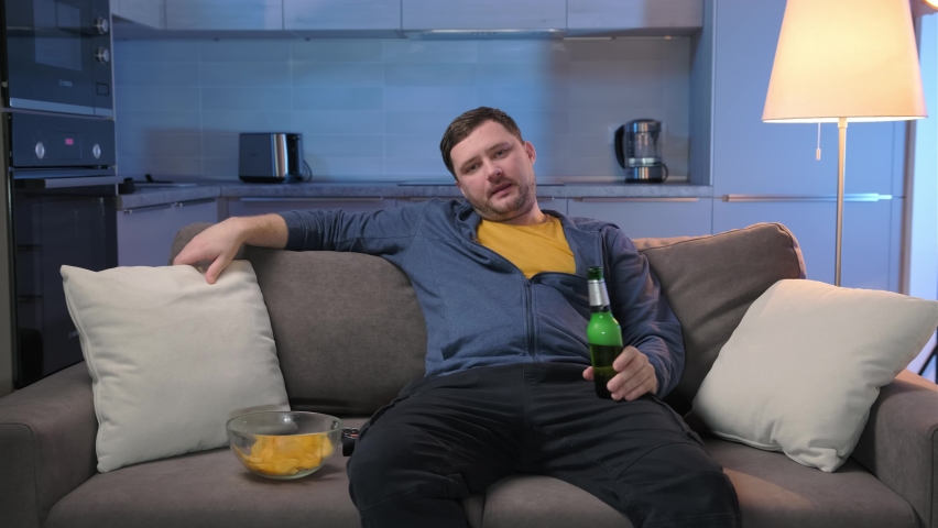 Single unshaven fat man suffering from insomnia switches TV channels on TV lying on couch and drinking alcoholic beer and potato chips on sofa in living room. Man spends evening at home in front TV. | Shutterstock HD Video #1067302246