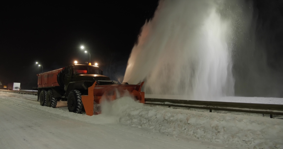 The grader removes snow on the road outside the city at night