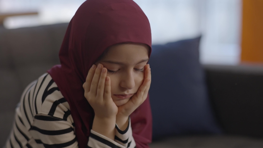 The Muslim little girl wearing a headscarf contemplates out of sorrow, holding her head in her hands. The sad little girl with a headscarf thinks deeply | Shutterstock HD Video #1067306488