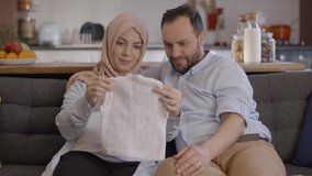 Muslim pregnant woman wearing a headscarf shows her husband the cardigan she knits for her future babies. Baby cardigan display concept. Slow motion video. - Powered by Shutterstock - Get 15% off with code: PIKWIZARD15