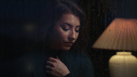 Attractive woman looking out from her home on a rainy night as water drips down her window and lightning flashes. Cinematic