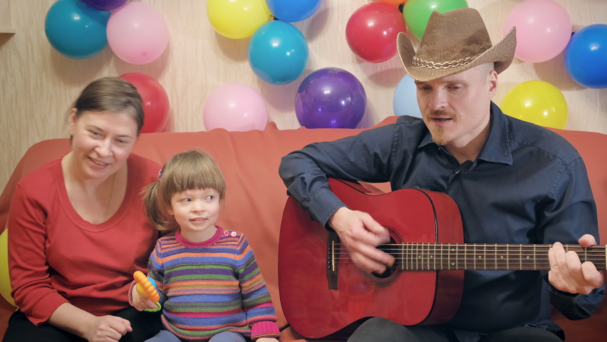 Father in cowboy hat plays guitar for his little daughter. Mother is sitting next to her and singing. There are lot of balloons around. Family is happy. Holiday, family weekend and birthday concept