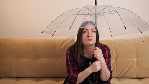 Sad brunette woman in checkered shirt hides from water flowing from upstairs neighbors under transparent umbrella sitting on sofa in room. Concept of flooding the apartment and property insurance