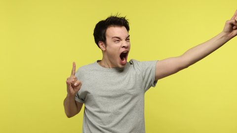 Delighted aggressive young man in gray t-shirt feeling crazy, showing rock and roll hand gesture and roaring, punk devil horns sign. Indoor studio shot isolated on yellow background