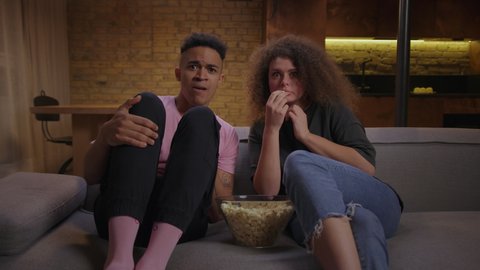 Millennial mixed race family watching scary movie sitting on couch with pop corn. Frightened man and woman watching TV and get scared suddenly.