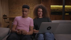 20s multiethnic couple talking to laptop sitting on couch at home. African American boyfriend with his girlfriend laughing together talking to friends by video call.