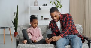 Young afro american father sits with his beloved daughter showing her video on phone, family watching horror movie on mobile device child punches hiding from fear, dad and girl kid spend time at home