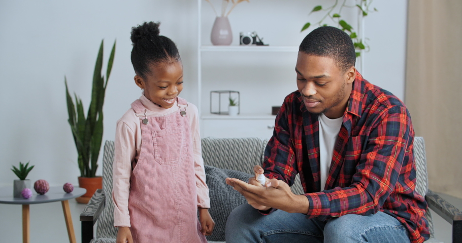 Afro american responsible father shows his little daughter example of good health care habit teaches how to use antibacterial gel sprinkles sanitizer on hands, dad and child treat palms from bacteria
