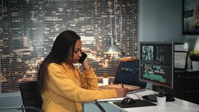 Mixed race colorist talking by smartphone and writing notes in modern production studio. In the backgrounds - skyscrapers.