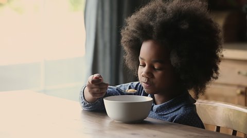 Head shot close up adorable small funny curly african american kid girl enjoying crunchy corn flakes with milk, eating morning fast breakfast with pleasure sitting alone at wooden table in kitchen.
