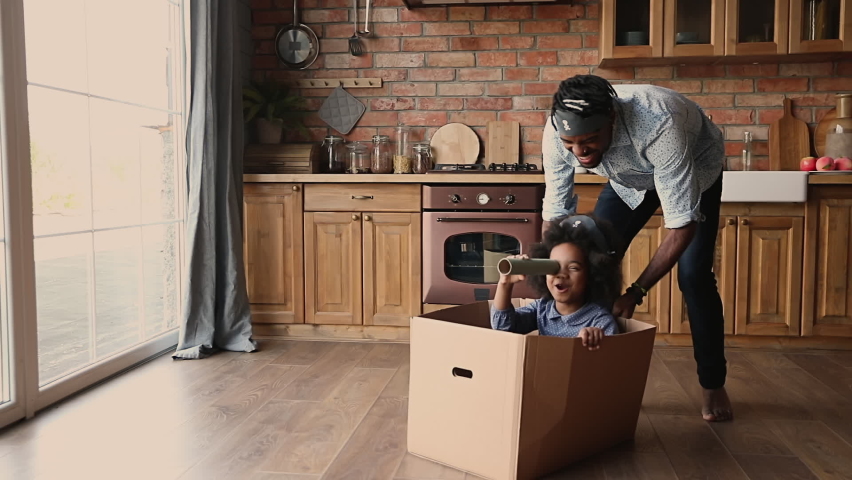 Joyful young african american father playing pirates with adorable small kid daughter sitting in box, imagining ship captain. Laughing little biracial child girl having fun with caring daddy indoors. | Shutterstock HD Video #1067335969