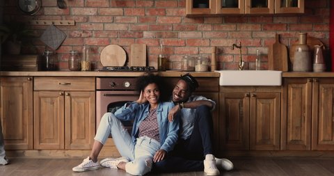 Full length romantic happy millennial african ethnicity family couple sitting on warm heated wooden floor in kitchen, dreaming of common future life, enjoying carefree weekend time in own house.