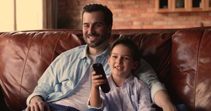 Affectionate smiling young dad cuddling happy joyful child son, watching smart TV programs, show, comedian movie or funny cartoons, enjoying free weekend leisure time resting on comfortable sofa.