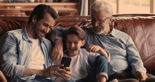 Smiling young man showing funny video on smartphone to laughing small boy son and sincere elderly mature father, relaxing together on comfortable couch, loving different generations family pastime.