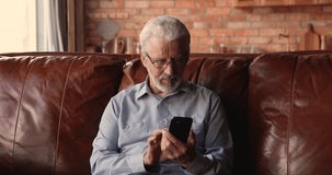 Happy middle aged 60s old man in glasses resting on comfortable sofa, involved in using cellphone applications, communicating distantly in social network, shopping, watching funny video photo content.