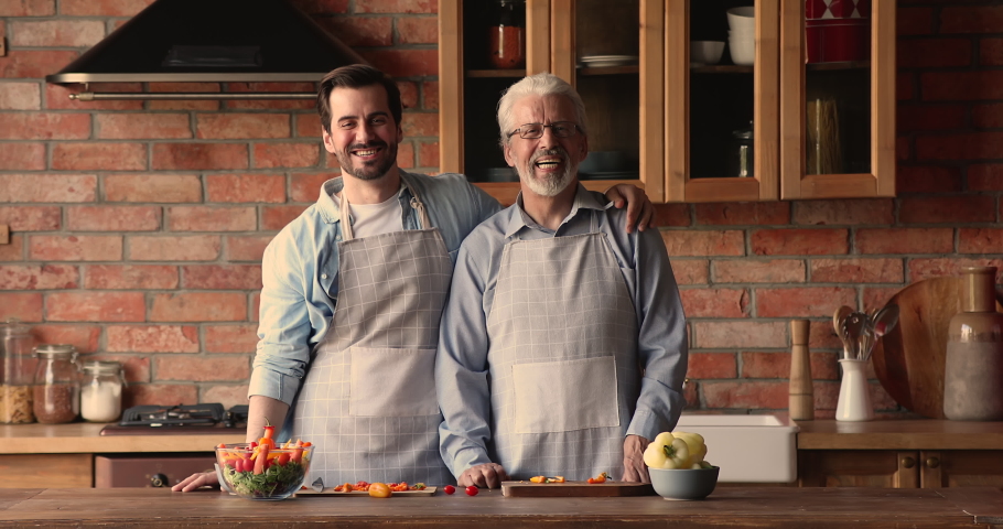 Happy bonding two male generations in aprons looking at camera, posing in old-fashioned kitchen. Smiling young man cuddling shoulders of sincere middle aged mature father, enjoying cooking together. Royalty-Free Stock Footage #1067337157