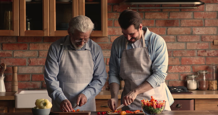 Happy two male generations family wearing aprons, enjoying cooking together in old-fashioned kitchen. Smiling old senior retired man preparing lunch with joyful grownup son on weekend at home. Royalty-Free Stock Footage #1067337166