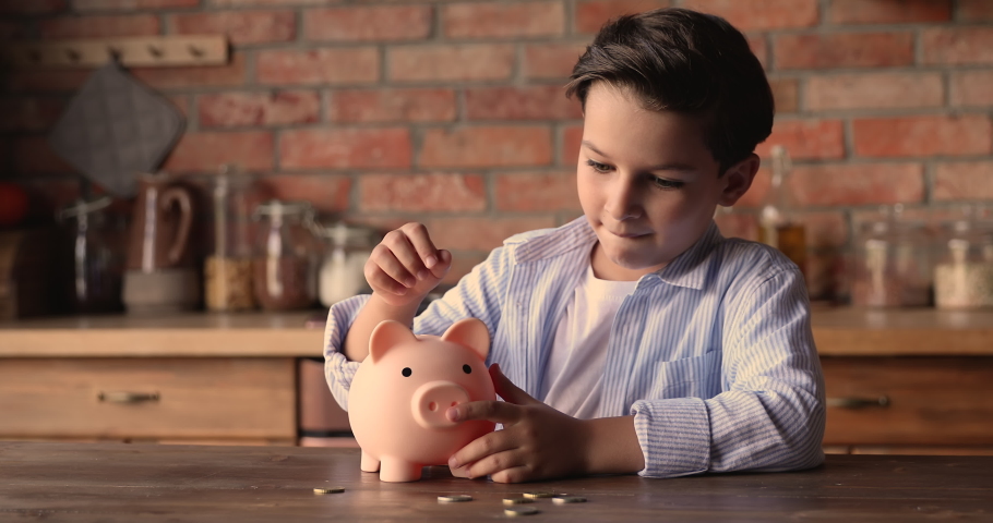 Happy small school boy putting coins into piggy bank, sitting at kitchen table at home, learning money savings for future, planning purchasing or shopping, financial literacy for children concept. Royalty-Free Stock Footage #1067337211