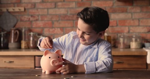 Happy small school boy putting coins into piggy bank, sitting at kitchen table at home, learning money savings for future, planning purchasing or shopping, financial literacy for children concept.