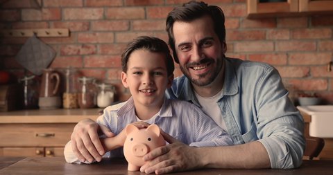 Happy young father cuddling small kid son, sitting at table in kitchen, holding piggybank in hands, future investment money savings, children financial literacy education, economy planning concept