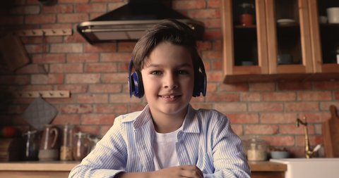 Adorable small 7s kid boy wearing wireless headphones, looking at camera, enjoying online class, holding video call meeting seminar training with teacher, distant education e-learning concept.