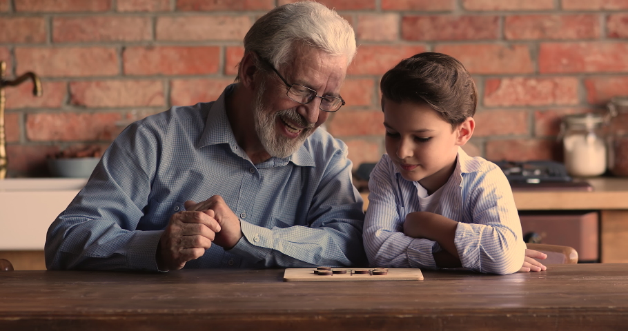 Happy elderly mature 60 hoary man in eyeglasses playing checkers draughts on wooden board with joyful little grandson, enjoying creative game on weekend leisure time, having fun together at home. Royalty-Free Stock Footage #1067337310
