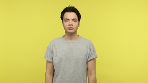 Young brunette man in casual style gray t-shirt holding breath with fingers on nose feeling unpleasant odor, disgusting aroma of emissions or fart. Indoor studio shot isolated on yellow background