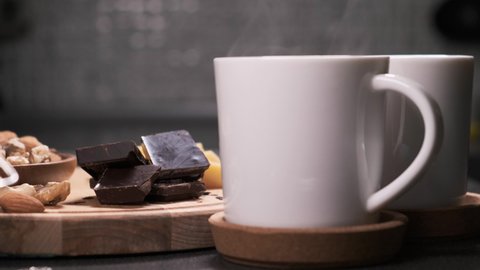Fresh coffee in two white mugs is on table and steam comes from it. woman or girl takes piece of bar of dark chocolate from the table.
