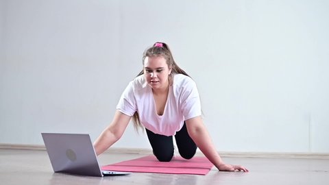 A young obese woman does push-ups. The girl is engaged in fitness at home on the mat and watches a training video on a laptop