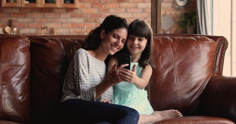 Young woman her little adorable daughter relaxing on sofa with smart phone making selfie photos having fun online, use new cool mobile application. Modern tech and offspring, parental control concept