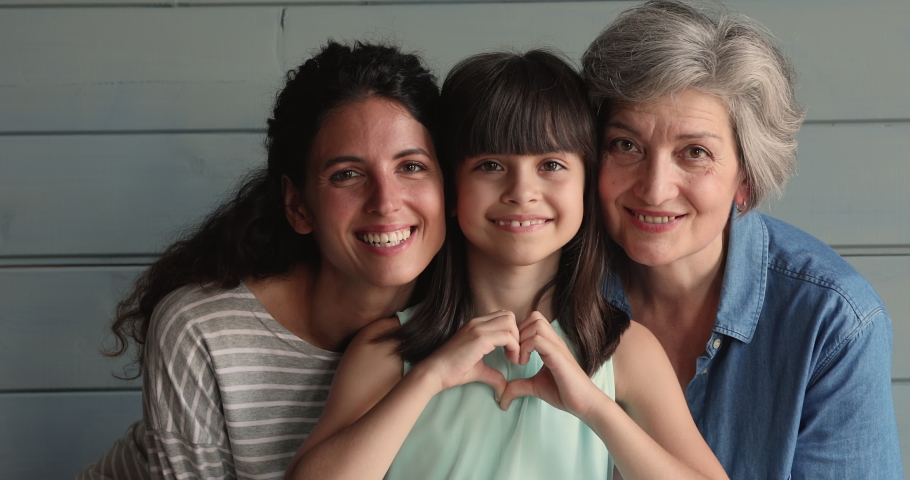 Little cute girl showing heart shape with fingers while pose in studio with elderly grandmother and mother smile look at camera. Three gen family portrait, life value, attachment and bonding concept