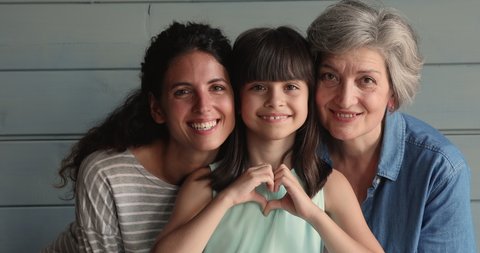 Little cute girl showing heart shape with fingers while pose in studio with elderly grandmother and mother smile look at camera. Three gen family portrait, life value, attachment and bonding concept