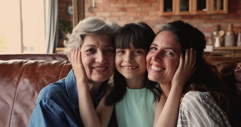 Grandma, mom and daughter of sit on sofa at home smile look at camera. Cute girl touch cheeks of mom and elderly granny enjoy moment of tenderness. Three generations of women family portrait concept