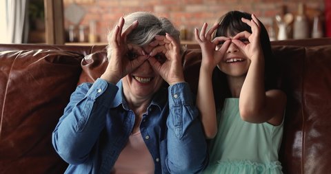 50s elderly granny her little 10s granddaughter sit on sofa making binocular shape with fingers look at camera through fake spectacles. Concept of having fun, eyesight vision care of older and younger