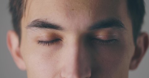 A close-up view of a calm handsome young man is opening his eyes and looking to the camera over grey background in slow motion