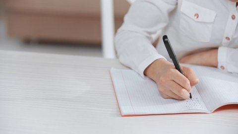 cropped view of schoolkid in white shirt writing in copy book, while doing homework at desk