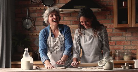 Elderly woman her grown up daughter preparing dough, flattening it with rolling-pin for bakery, talk laughing enjoy cookery and communication in cozy kitchen. Two generation women family hobby concept