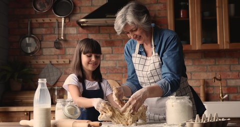 Elderly 60s grandmother preparing with little pretty granddaughter family recipe pie, kneading with hands dough talking enjoy process. Older generation teach younger, hobby and cookery at home concept