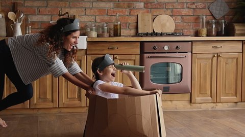 Young mother ride little cute daughter sit in carton box, kid girl looking in paper tube binocular enjoy creative game with mom in kitchen. Creativity, handmade plaything, having fun at home concept